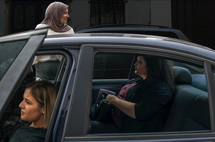 Två kvinnor sitter i en bil och en kvinna står utanför. Bildörren är öppen.