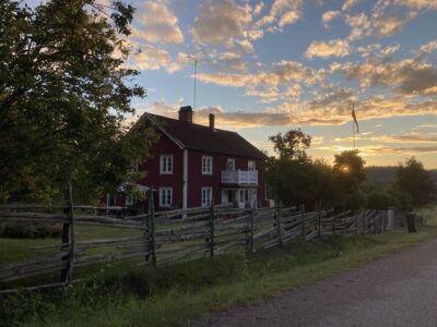 Ett rött hus på landet med ett staket framför. Solnedgång i bakgrunden.
