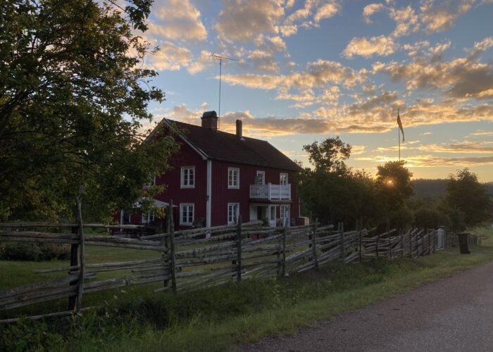 Ett rött hus på landet med ett staket framför. Solnedgång i bakgrunden.