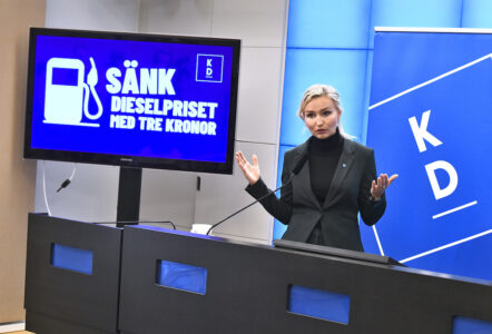 KD-ledaren Ebba Busch talar framför en skärm med bild på en bränsledunk och texten "Sänk dieselpriset med tre kronor".