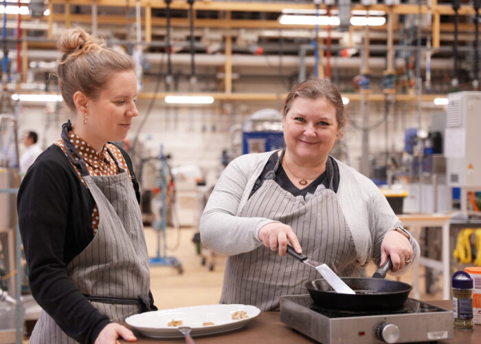 En forskare steker något i en stekpanna i ett stort provkök, ler och tittar in i kameran. En annan forskare betraktar den första.