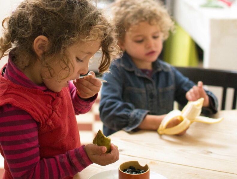 Två barn sitter vid ett bord och äter päron respektive banan.
