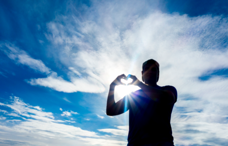 Man formar ett hjärta med sina händer, i bakgrunden blå himmel och vita moln.