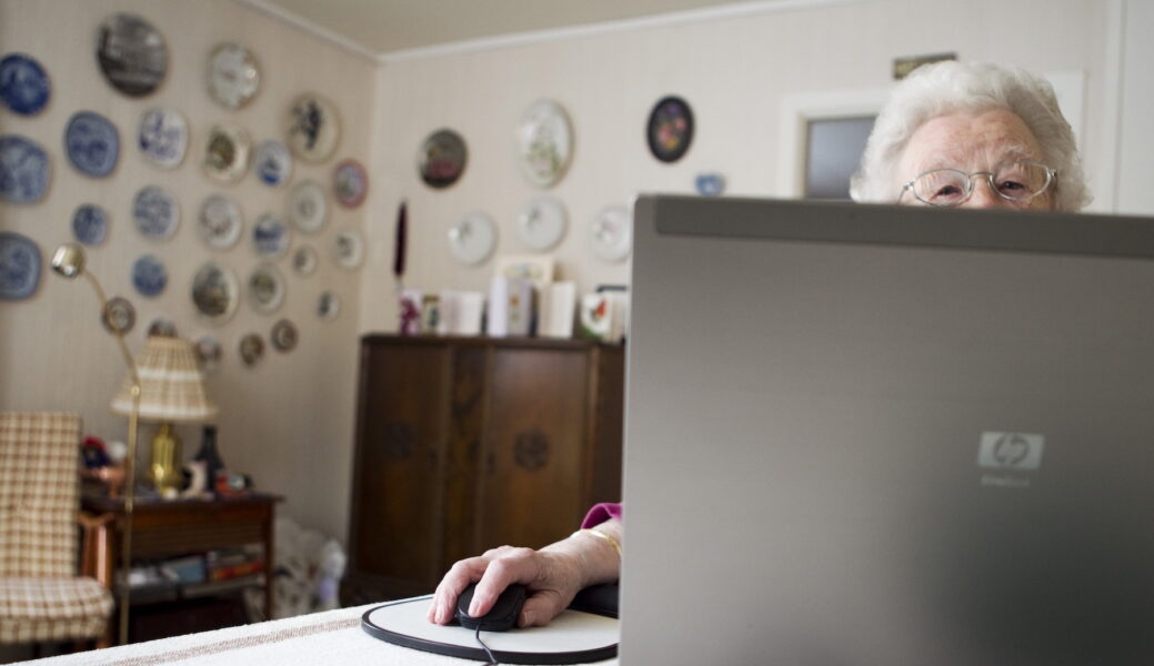 Äldre kvinna i hemmiljö, halvt dold bakom en laptopskärm.