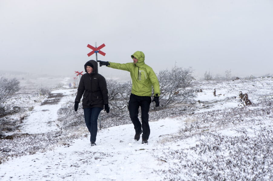 Två vandrare längs en utmarkerad led i snölandskap, den ena pekar ut ur bild och den andra tittar dit.