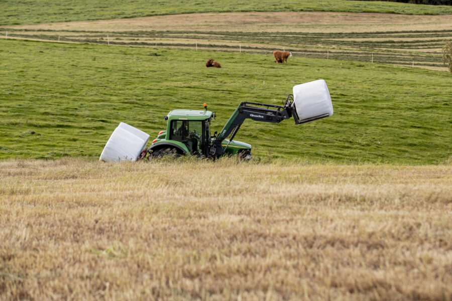 En traktor kör över en åker, lastad med två plastinklädda rundbalar.