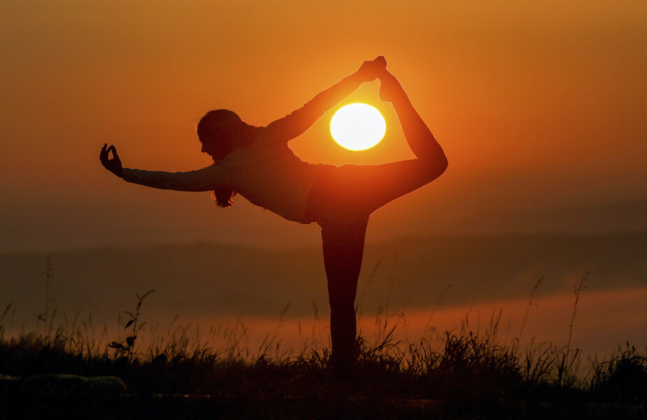 Silhuett av person som står på ett ben och håller i sin fot bakom huvudet, i bakgrunden en solnedgång.