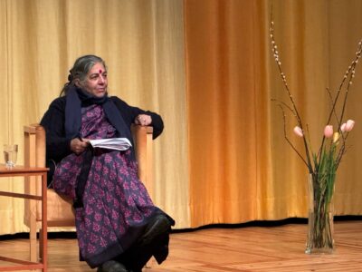Vandana Shiva klädd i lila sitter på en scen framför ett gult draperi, med en bukett blommor till höger i bild.