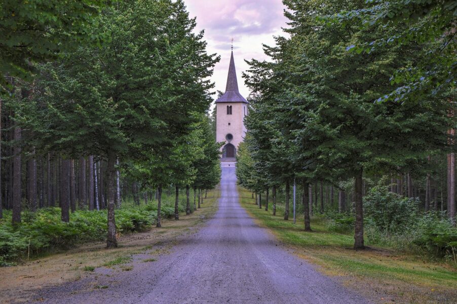 Vit kyrka med spetsigt tak syns i mitten av bilden, en rak väg kantad av skog leder dit.