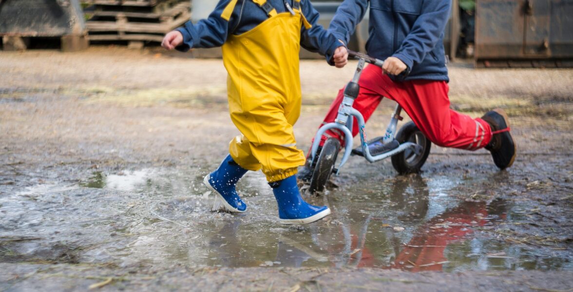 Två barn i regnkläder springer respektive cyklar i en vattenpöl. Ansiktena syns ej.