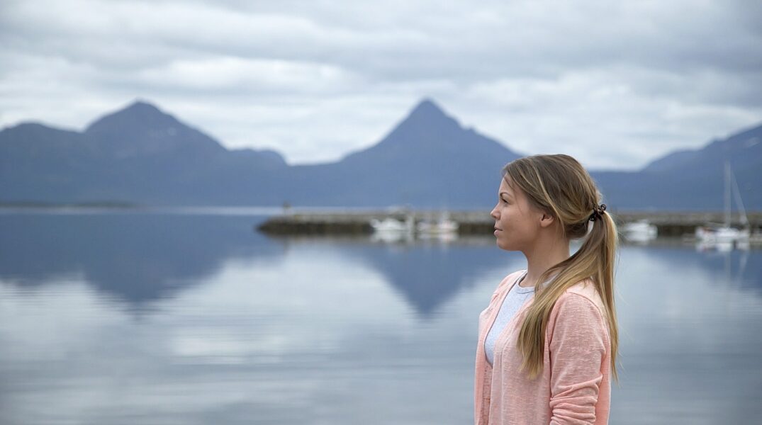 Blond kvinna i profil med en sjö och berg i bakgrunden.