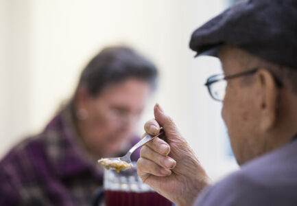 Äldre man i färd att ta en tugga mat med en sked. I bakgrunden en annan äldre person.