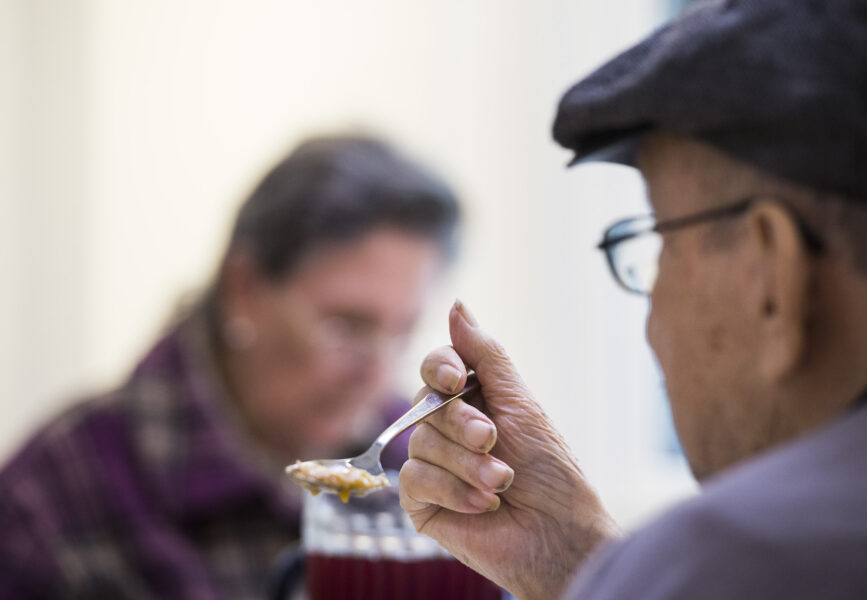 Äldre man i färd att ta en tugga mat med en sked. I bakgrunden en annan äldre person.