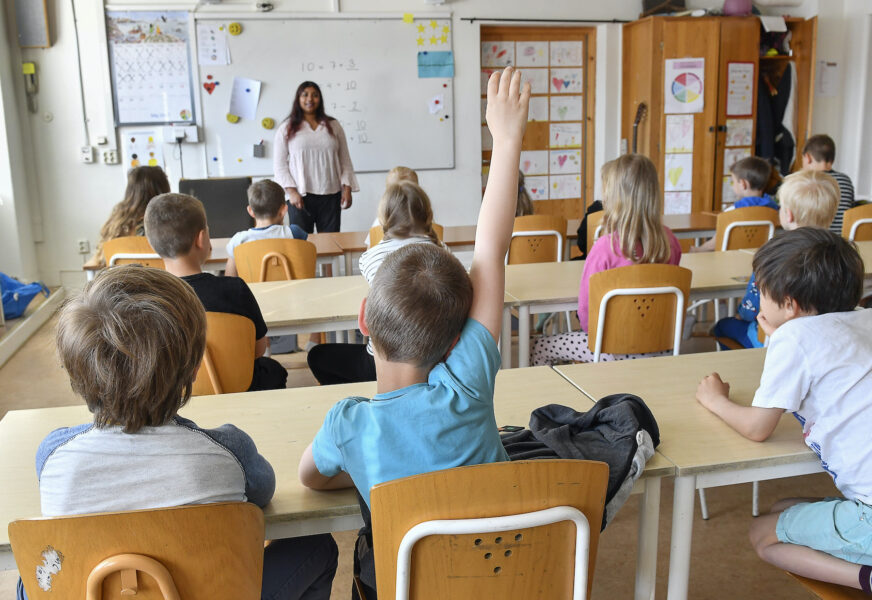 Klassrum sett från elevens synvinkel. En elev räcker upp handen. I bakgrunden en lärare framme vid whiteboardtavlan.