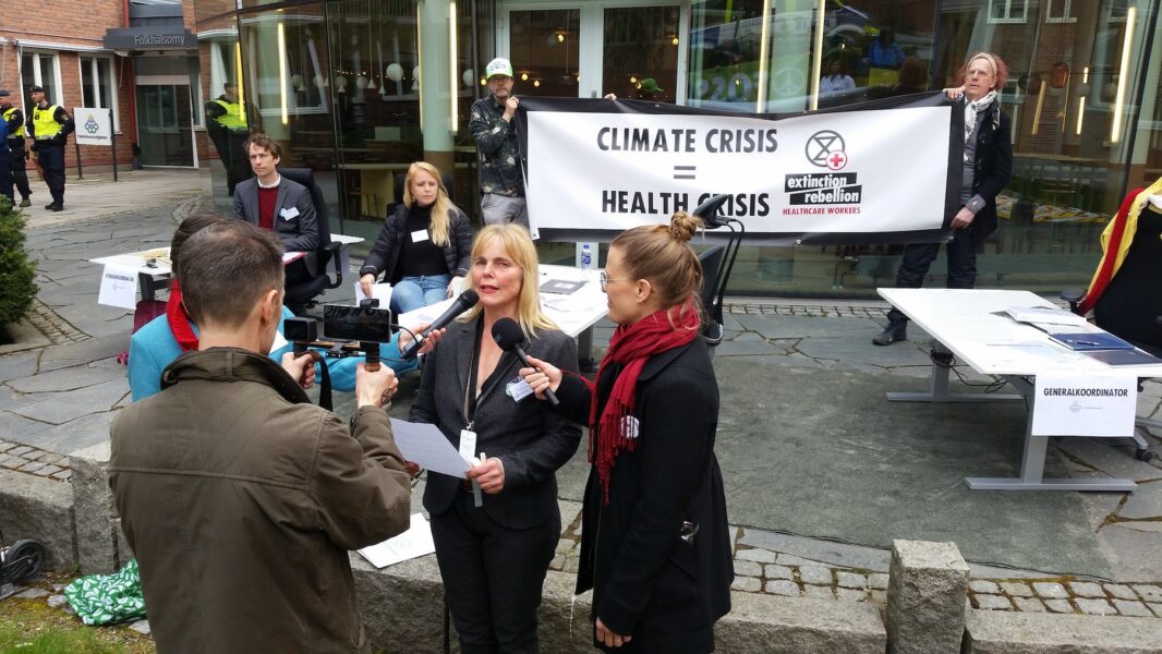 Aktivister med kontorsbord och stolar utanför Folkhälsomyndigheten håller upp ett plakat med texten Climat crises=Health crises
