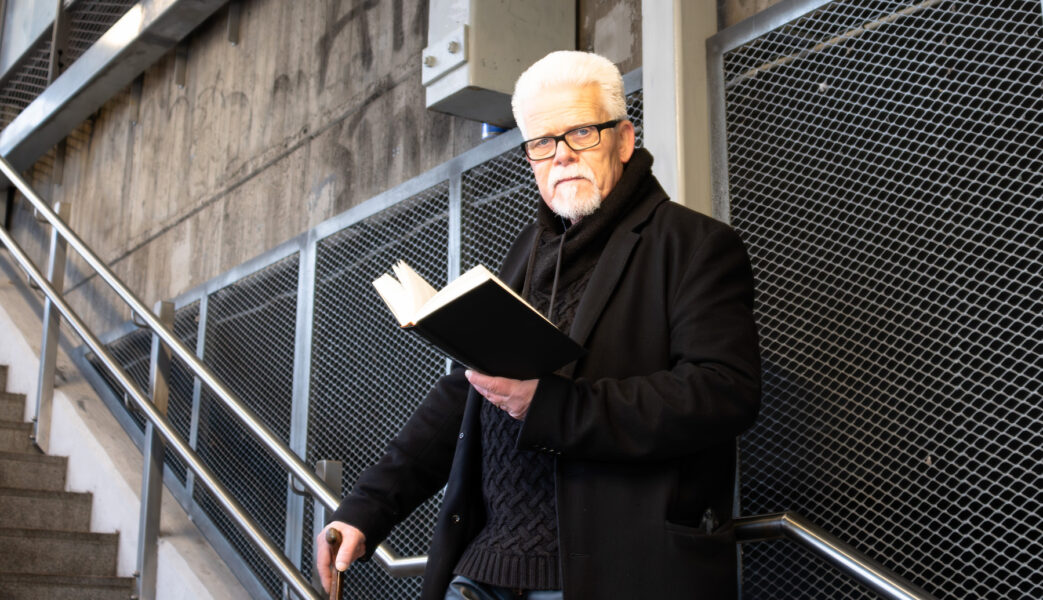 Mattias Gardell står i en trappa och tittar in i kameran, svartklädd med en uppslagen svart bok i enda handen och en käpp i andra.