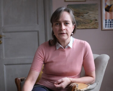 Porträtt av Nina Wormbs, rosaklädd i en sliten grå fåtölj.