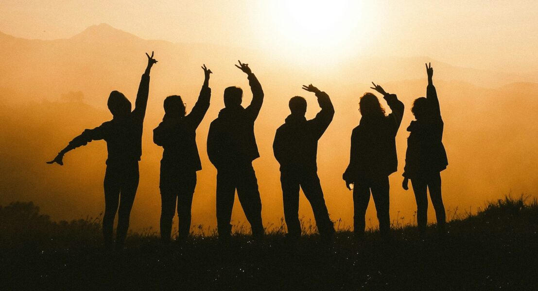 Sex personer i silhuett mot en solnedgång, några gör segertecken med händerna.