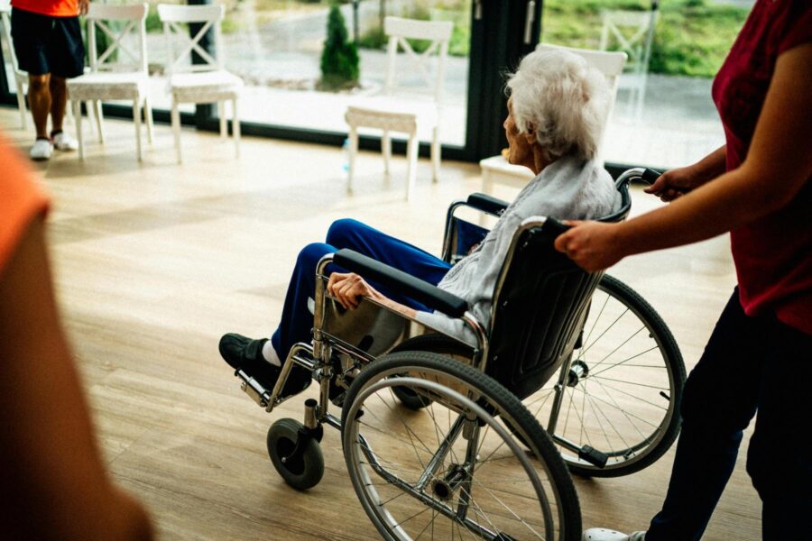 Äldre kvinna körs i rullstol av en yngre kvinna på ett vårdhem.