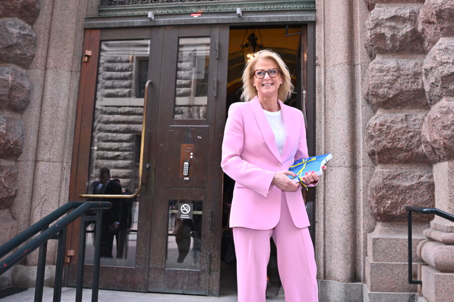 Elisabeth Svantesson i rosa kostym, leende utanför en port, med vårändringsbudgeten i händerna.