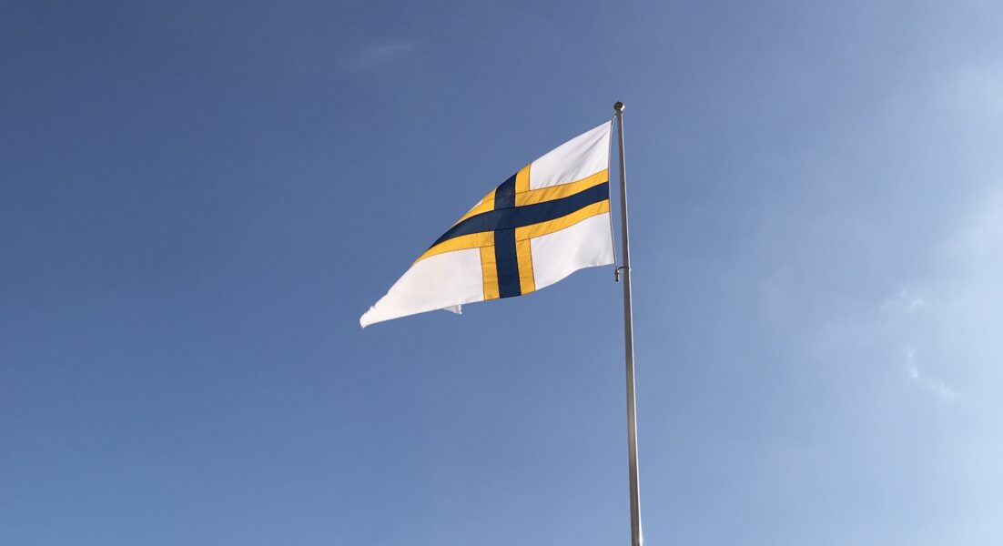 Sverigefinska flaggan, vit med ett blågult kors, vajar i vinden mot blå himmel.