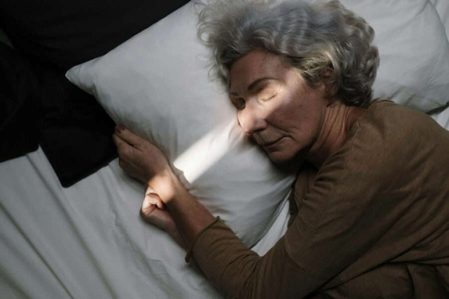 Äldre kvinna med grått hår och brun tröja ligger på en säng med kudde.