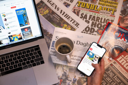 Kaffekopp står på flera dagstidningar invid en datorskärm och mobilskärm.