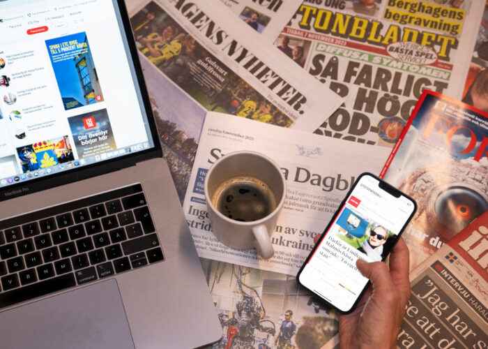 Kaffekopp står på flera dagstidningar invid en datorskärm och mobilskärm.