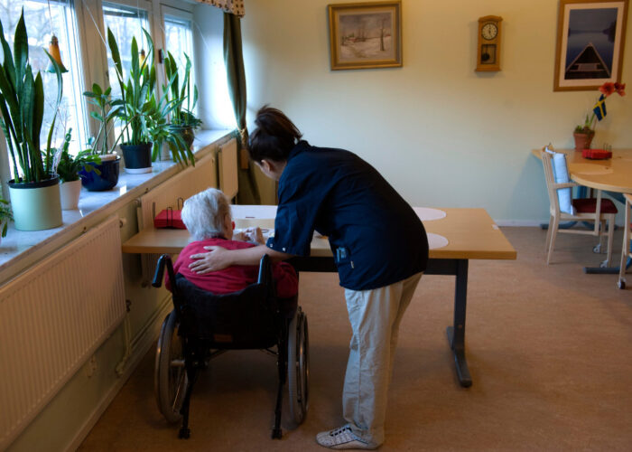 En äldre person sitter i rullstol vid ett bord, en sjukvårdare håller en hand på hens rygg.