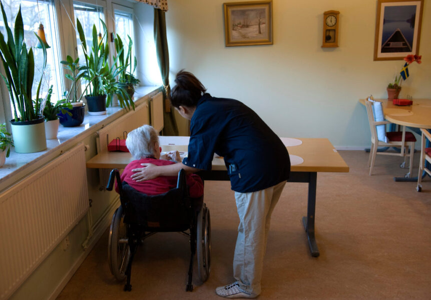 En äldre person sitter i rullstol vid ett bord, en sjukvårdare håller en hand på hens rygg.