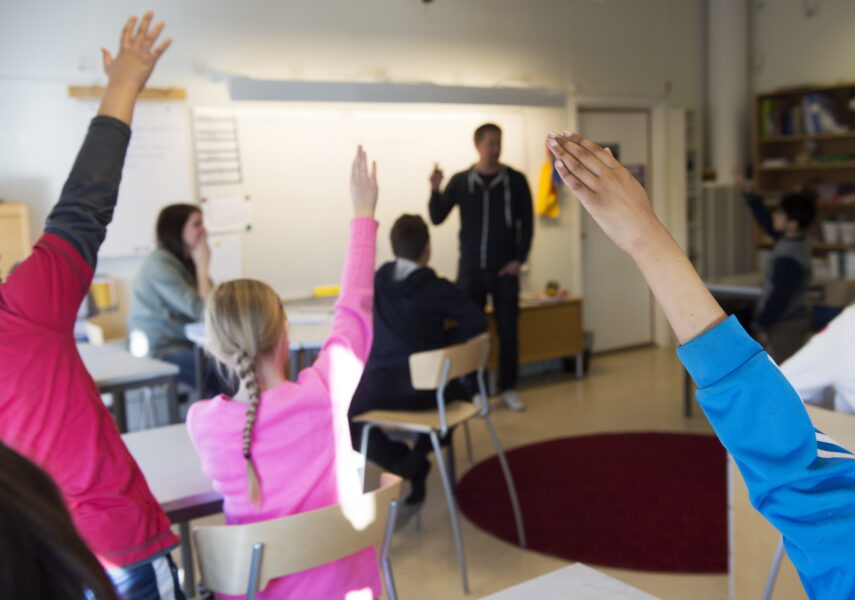 Skolklass där eleverna räcker upp handen, längst fram en lärare vid en whiteboard.
